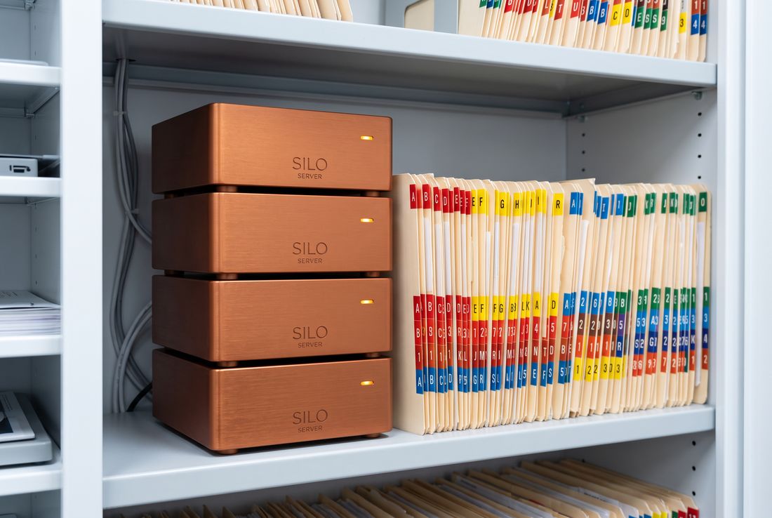 Four Silo Server modules stacked in a column on a shelf inside a clinical records cabinet, beside colour-tabbed patient file folders